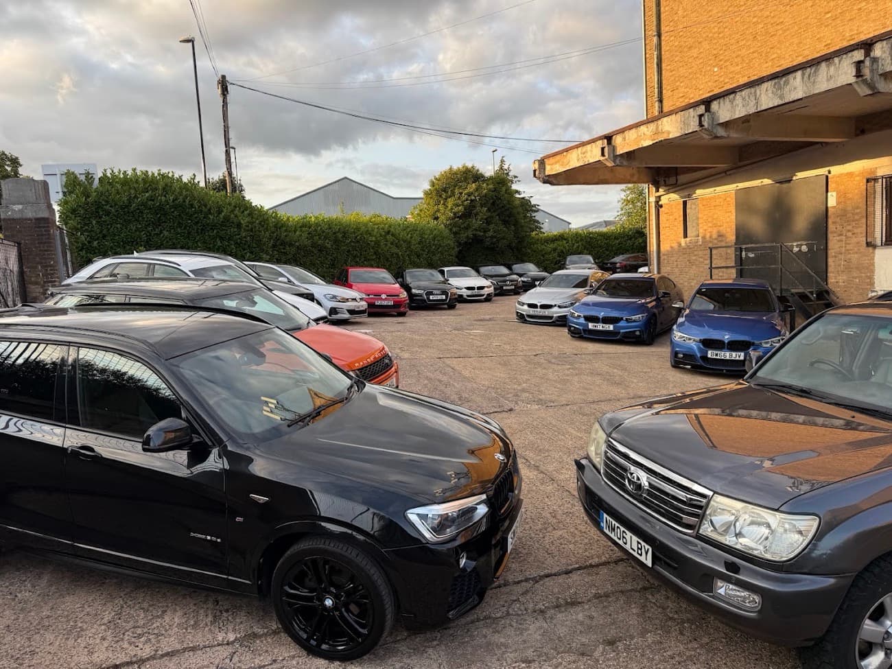 Before: a crowded used car yard at dusk, a black BMW SUV in the left foreground among a dozen other cars, a hedge and a brick building