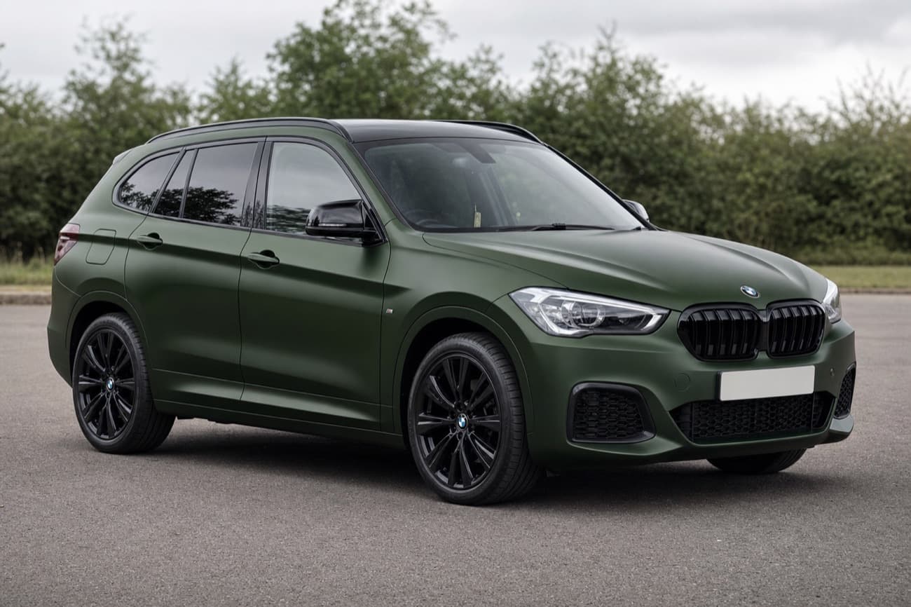 A BMW SUV in a satin forest green wrap with black wheels, parked on tarmac in front of trees