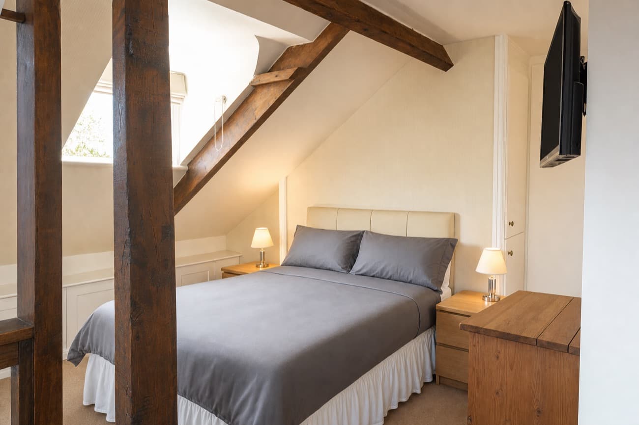 After: the same attic bedroom in landscape, the same beams, bed and furniture, the bed made, the surfaces clear and both lamps on