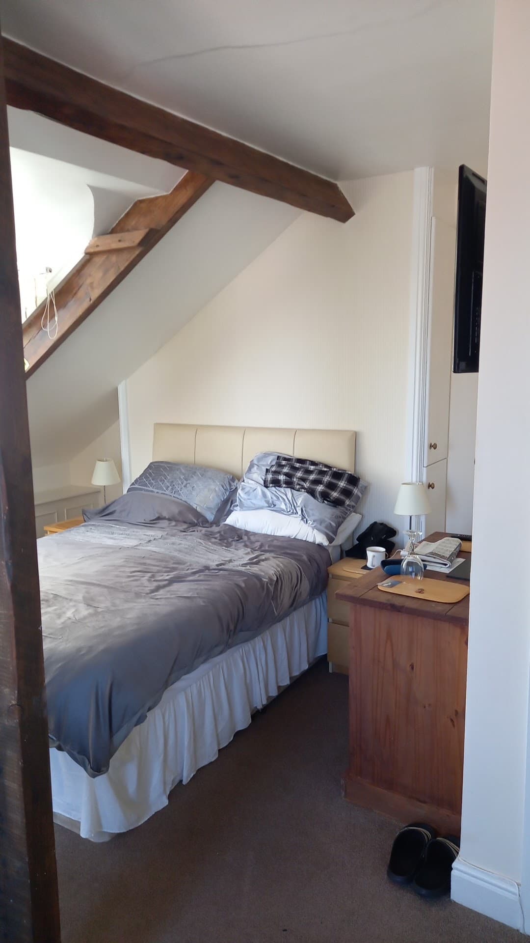 Before: a portrait phone photo of an attic bedroom from the doorway, the bed unmade, a mug, glasses and clutter on the bedside table, shoes on the floor