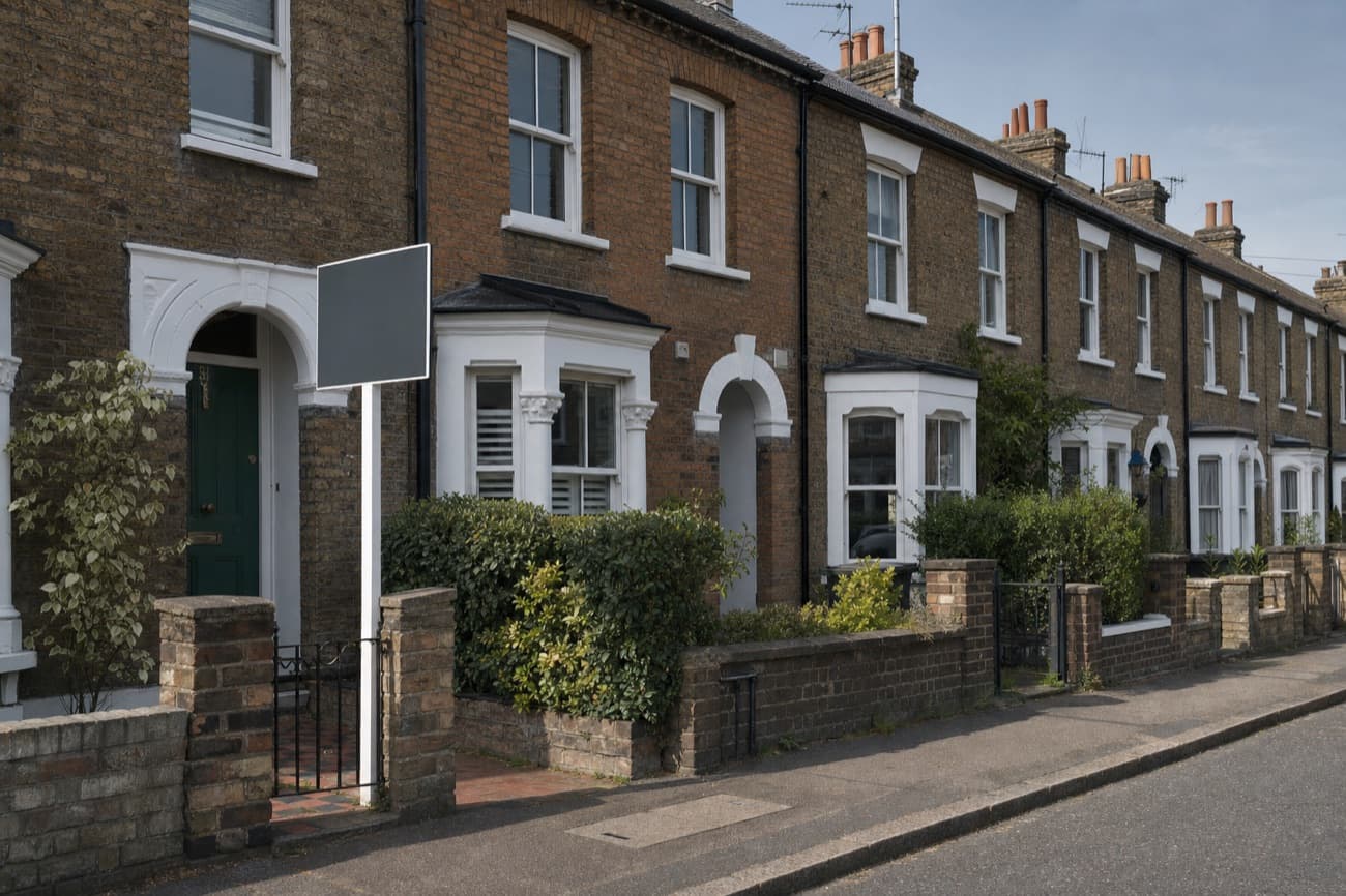 A row of brick terraced houses with white bay windows, a blank estate agent's board on a post by the first front gate
