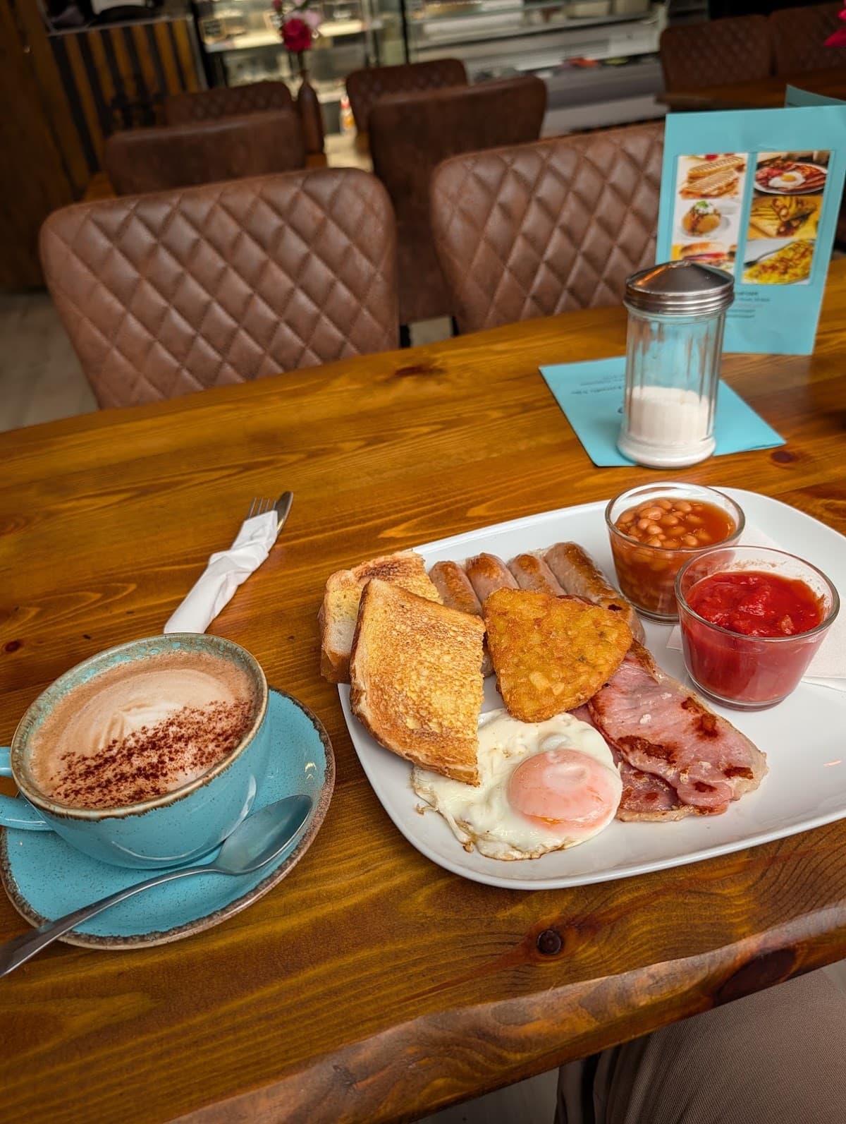 Before: a customer's phone photo of a full English breakfast on a white plate, shot from above at a tilt, with a blue cappuccino cup and a lot of bare wooden table