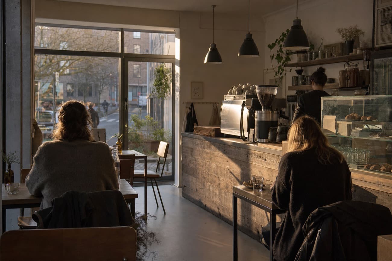 A sunlit cafe in late afternoon, two customers seated with their backs to the camera, a wooden counter and a cake case beyond them