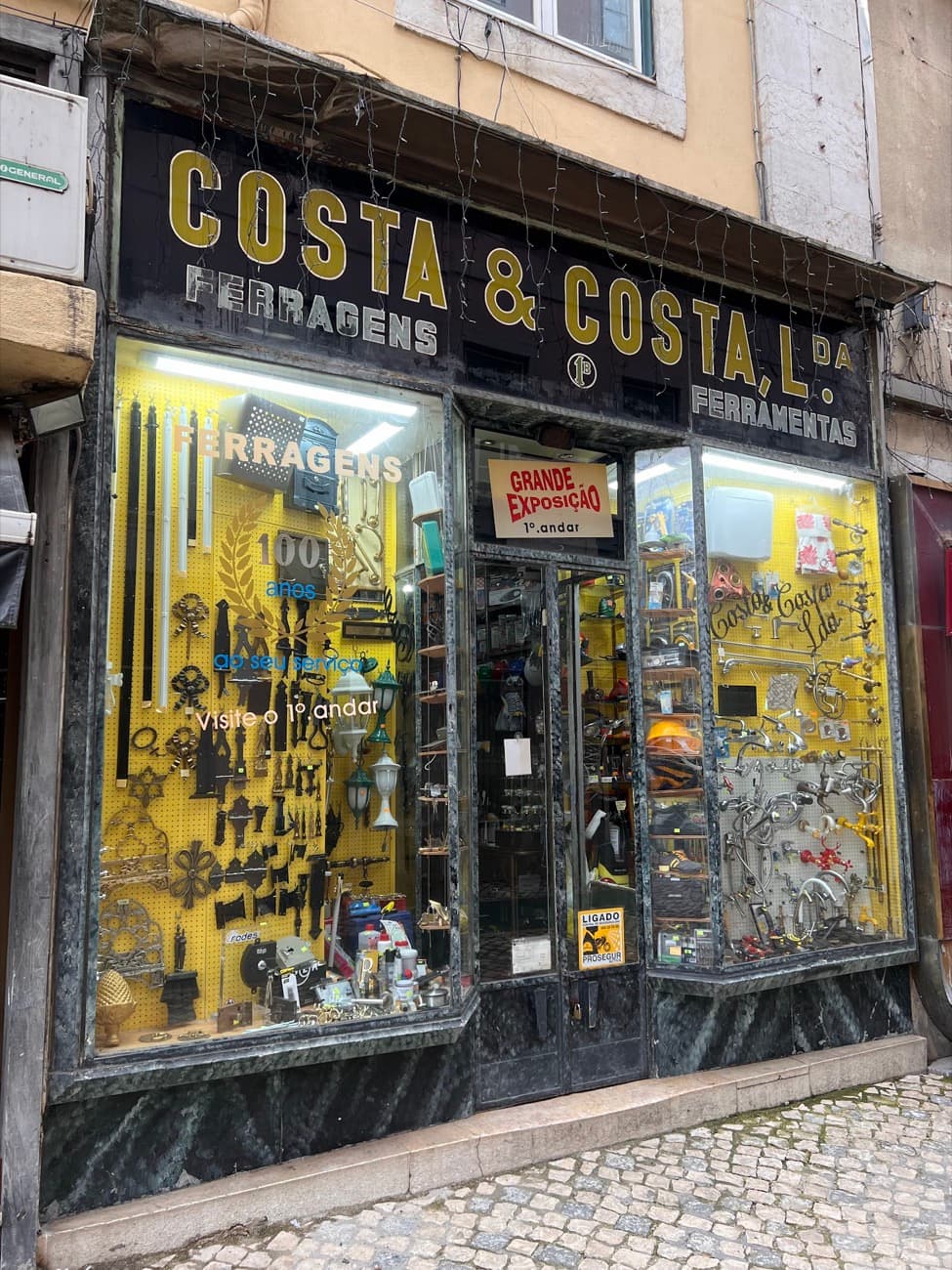 Before: Costa & Costa's frontage on a grey day, black and gold fascia, yellow pegboard windows, dead wiring across the sign